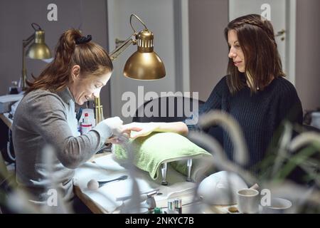 Felice estetista facendo manicure a un cliente sorridente sotto la lampada d'oro nel salone delle unghie. Donna ottenere manicure unghie. Foto Stock