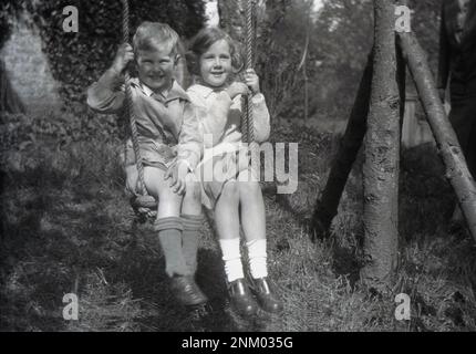 1932, storico, estivo e all'aperto in un giardino selvaggio, un giovane ragazzo e una ragazza, fratello e sorella, che si divertono seduti accanto l'uno all'altro sulla sede di un'oscillazione fatta a mano in corda da giardino, Inghilterra, Regno Unito. Foto Stock