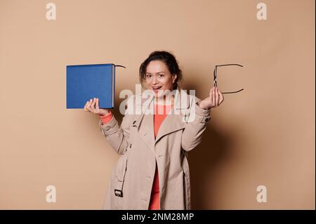Felice insegnante femminile di mezza età positiva in abiti casual beige, posa con un libro e occhiali su sfondo crema Foto Stock