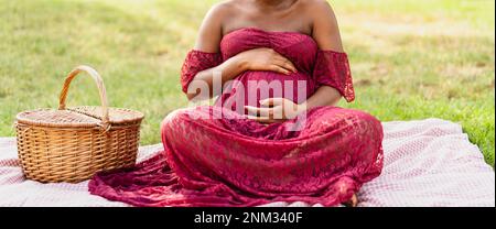 Donna africana carezzando il suo ventre incinta mentre fa un picnic nel parco - concetto di stile di vita di maternità Foto Stock