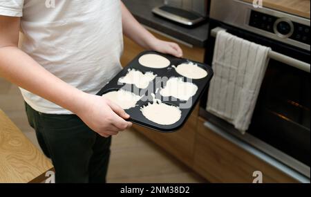 vassoio per bambini in sovrappeso con cupcake crudi, muffin, pasta. Persona senza volto in piedi vicino al forno in una moderna cucina interna. Festa delle madri Foto Stock