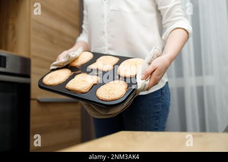 Donna che tiene vassoio caldo con cupcake al forno, muffin, biscotti in potholder, in piedi vicino al forno in cucina moderna. Cucina fatta in casa. Persona senza volto Foto Stock