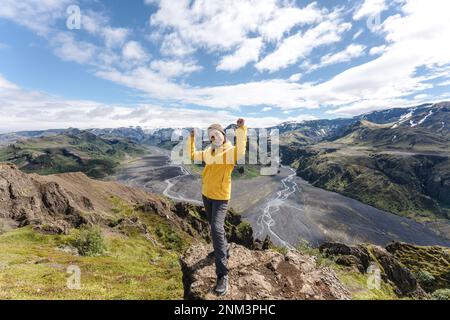 Successo di escursionista femminile in giacca gialla in piedi e sorridente in cima alla montagna in Valahnukur punto di vista in estate a Thorsmork, Islanda Foto Stock