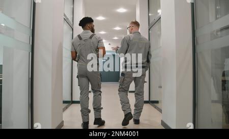 Vista posteriore di due uomini diversi in uniforme corridoio a piedi e discutere telecamere CCTV o l'installazione di Internet in ufficio coworking. I programmi di installazione configurano le telecamere utilizzando un computer portatile. Concetto di sistema di sorveglianza. Foto Stock