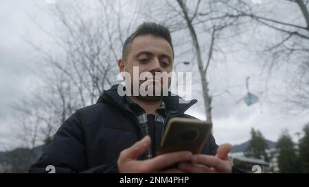 Giovane uomo con pelle giusta e capelli corti sorridente mentre utilizza il suo smartphone per Texting o fare pagamenti online all'aperto Foto Stock