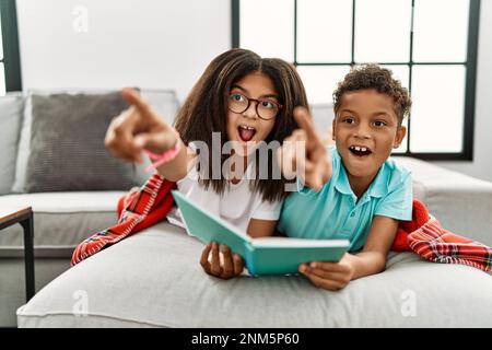 Due fratelli sdraiati sul divano leggendo un libro che punta con il dito sorpreso davanti, bocca aperta stupita espressione, qualcosa sulla parte anteriore Foto Stock