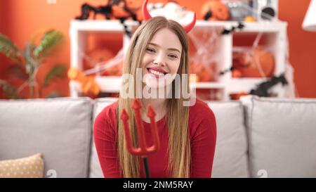 Giovane donna bionda sorridente indossando il costume diavolo seduto sul divano a casa Foto Stock