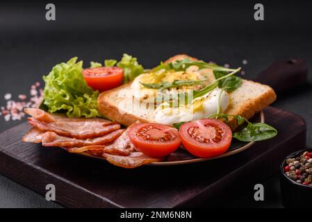 Delizioso pranzo salutare composto da pancetta, pane tostato, uova, pomodori, lattuga e verdure su un piatto nero su uno sfondo di cemento Foto Stock