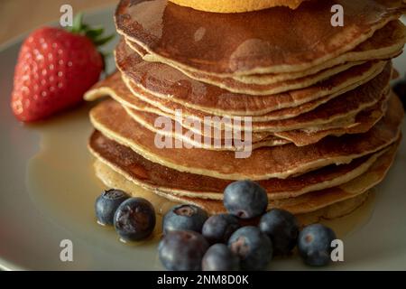 Primo piano di deliziosi pancake su un piatto con sciroppo, mirtilli, una fragola e una fetta d'arancia in un ambiente ben illuminato Foto Stock