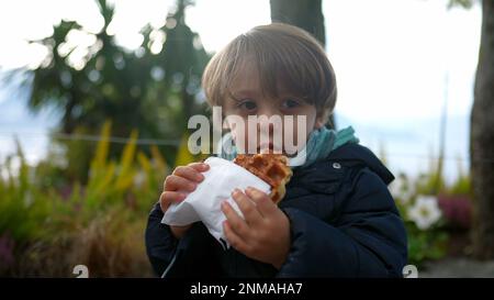 Un ragazzino felice che mangia waffle belgi all'aperto. Ragazzo carino ridendo e sorridendo mentre mangiava cibo di strada Foto Stock