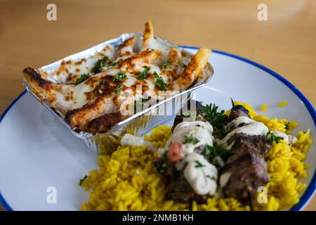 Un kabab di manzo greco mediterraneo su riso allo zafferano su un piatto con un lato di patatine fritte greche ricoperte di mozzarella fusa e formaggio feta, erbe aromatiche, e. Foto Stock