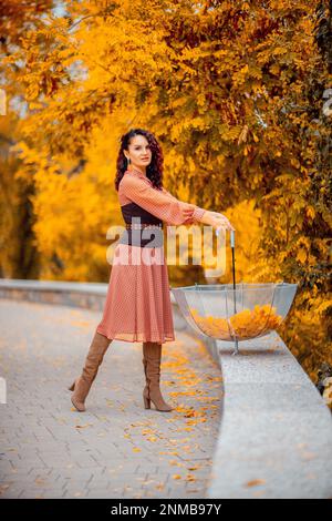 Bella ragazza in un vestito con un ombrello nel parco autunnale. Lei lo tiene sopra la testa, foglie autunnali stanno cadendo da lui Foto Stock