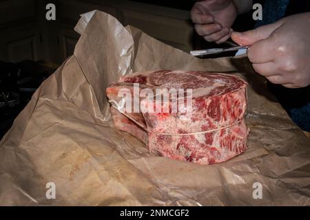 Crudi due ossa stnading costola arrosto di manzo su carta macellaio con pepe su di essa essendo spruzzato con sale da un cucchiaio di rombo argento a mano Foto Stock