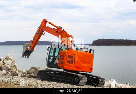 Nuovo retroescavatore arancione brillante su piste da lago che dispone grandi massi vicino all'acqua Foto Stock