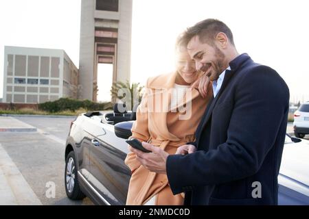Allegro coppia di mezza età guardando telefono cellulare appoggiato su auto convertibile. Ritratto moglie e marito Foto Stock
