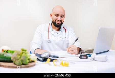 Sorridente scrittura nutrizionista in ufficio. Scrittura nutrizionista su blocco note. Nutrizionista maschile che lavora con appunti e computer portatile alla scrivania Foto Stock