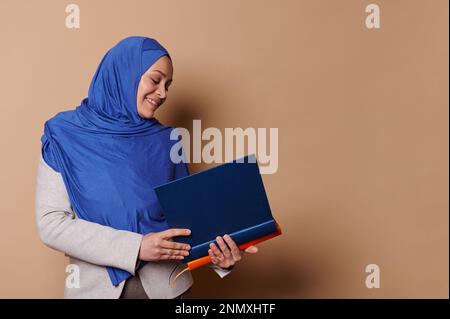 Donna musulmana fiduciosa in hijab, sorridente, tenendo un mucchio di libri di copertina rigida nelle sue mani, isolato su sfondo beige Foto Stock