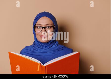 Primo piano ritratto sorridente donna musulmana in hijab blu, legge libro e sorride alla macchina fotografica, sfondo beige. Persone. Istruzione Foto Stock