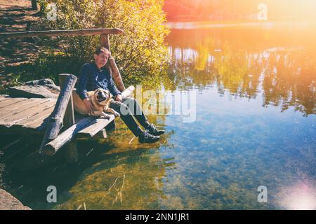 Uomo con il cane Labrador Retriever seduto su un ponte di legno presso la riva del lago e guardando l'acqua Foto Stock