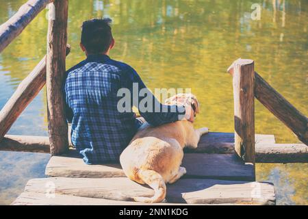 Uomo con il cane Labrador Retriever seduto su un ponte di legno presso la riva del lago e guardando l'acqua Foto Stock