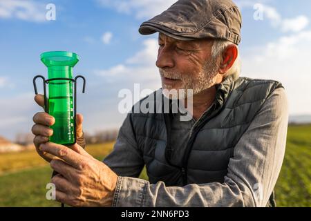 Agricoltore anziano che guarda il misuratore di acqua per misurare la pioggia in campo o giardino Foto Stock