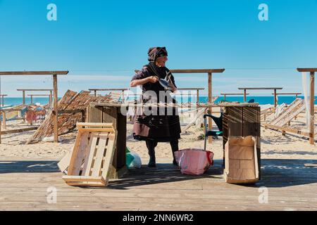 Nazare, Portogallo - 9 dicembre 2022 : vecchia signora in nero vende pesce secco sulla spiaggia del villaggio di pescatori sulla costa atlantica Foto Stock