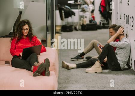 Colleghi maschi seduti sul pavimento e con una conversazione mentre la loro collega femminile dai capelli ricci si trova su un divano rosa e lavora in grembo Foto Stock