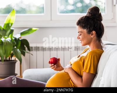 Bella giovane donna che si prepara a mangiare mela rossa sul divano a casa. Stile di vita sano durante il periodo di maternità Foto Stock