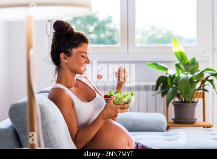 Bella giovane donna che mangia insalata sul divano a casa. Stile di vita sano durante il periodo di maternità Foto Stock
