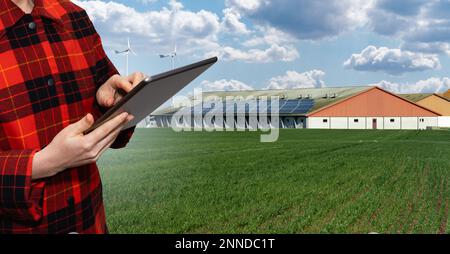 Agricoltore con computer tablet sullo sfondo di una moderna fattoria casearia che utilizza energia rinnovabile, pannelli solari e turbine eoliche Foto Stock