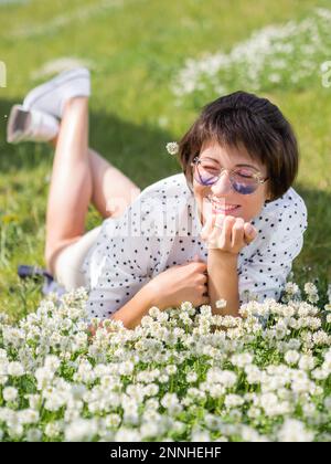 Donna in occhiali da sole colorati sniffs fiori trifoglio sul prato in parco urbano. Natura in città. Rilassati all'aperto dopo il lavoro. Atmosfera estiva. Foto Stock