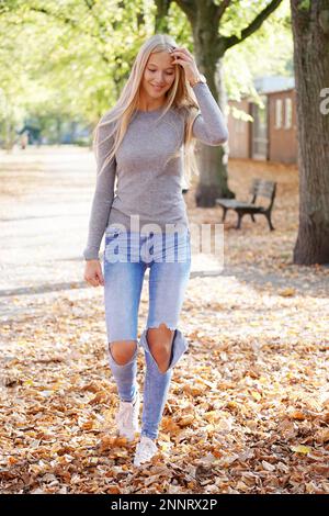 adolescente ragazza godendo a piedi in un parco in una giornata di sole in autunno - giovane donna bionda che indossa il maglione e jeans stressati alla moda Foto Stock