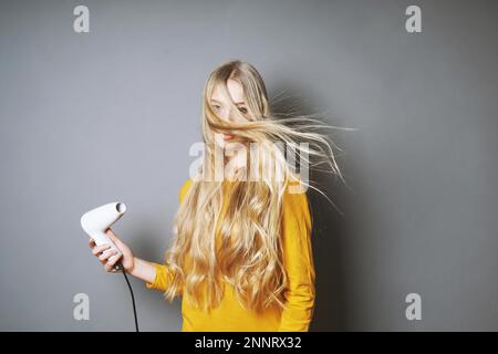 Funny giovane donna bionda asciugare i suoi lunghi capelli con phon Foto Stock