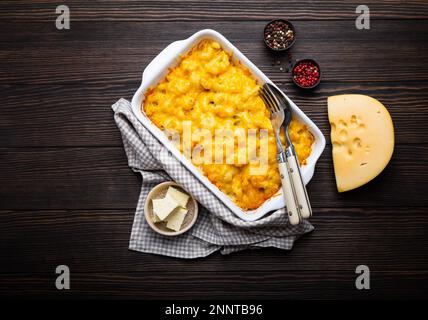 Primo piano di mac al forno e formaggio in casseruola bianca su sfondo rustico in legno, con condimenti, burro, cucchiaio e forchetta, vista dall'alto. Pasta con formaggio Foto Stock