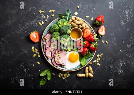 Ketogenic basso concetto di dieta dei carbs, vista dall'alto. Piatto su fondo nero di pietra con cibi keto: Uova, carne, olio d'oliva, broccoli, bacche, noci, semi Foto Stock