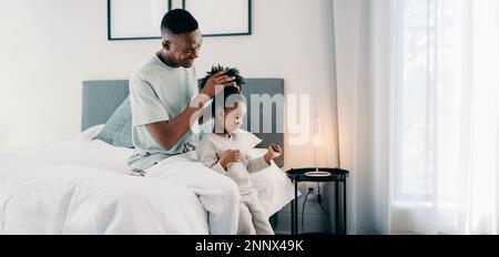 Il papà nero piega i capelli di sua figlia Afro in un panino mentre si siede su un letto. Padre dona a sua figlia una nuova acconciatura. Papà e figlia che spendono il Foto Stock