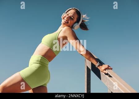Donna felice in piedi contro il cielo in abbigliamento sportivo. Donna sportiva che ama ascoltare musica con le cuffie. Donna che lavora fuori al sole del mattino. Foto Stock