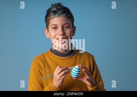 Felice ragazzo caucasico di 10 anni con capelli blu che ricoprono un occhio con un uovo di Pasqua decorato su sfondo blu. Perfetto per design a tema pasquale o. Foto Stock