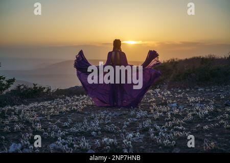 Tramonto viola abito donna montagne. Ascesa del mistico. tramonto sulle nuvole con una ragazza in un lungo vestito viola. Nel prato c'è un prato Foto Stock