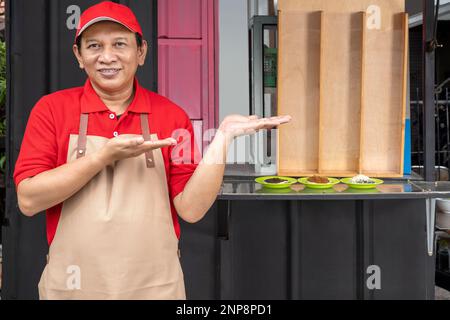 Uomo asiatico con cappello e grembiule con palma aperta che mostra qualcosa di fronte al negozio. Serabi è una tradizionale frittella indonesiana Foto Stock