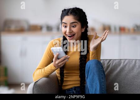 Giovane donna indiana entusiasta guardando lo schermo dello smartphone a casa Foto Stock