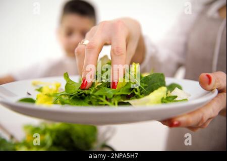 cottura insalata mamma con mano aggiunge le foglie di lattuga a grande ciotola in background ragazzo agita insalata cucina cottura grembiuli cottura casalinga cibo comunicazione bambini aiutare il bambino in cucina formazione Foto Stock