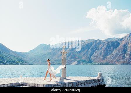 Sposa in un abito che fruleggia nel vento cammina lungo il vecchio molo oltre la colonna Foto Stock