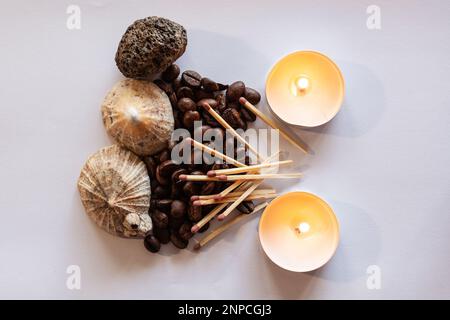 Un mucchio di chicchi di caffè è circondato da conchiglie e da una pietra vulcanica. I fiammiferi sono posti sui chicchi di caffè e due candele sono sul lato. Foto Stock