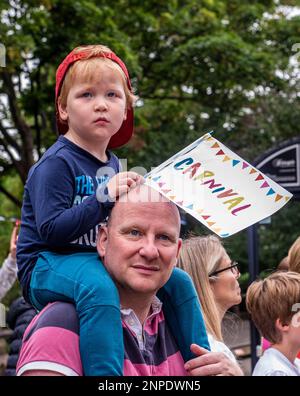 Un ragazzo si siede sulle spalle del papà con una bandiera di Carnevale e guarda alla sfilata dei costumi del Carnevale di Harrogate. Foto Stock