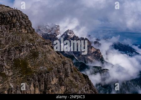 Scogliere rocciose e cime dei monti Zwölferkofel e cima d’Auronzo, parzialmente avvolti da nuvole, due escursionisti in piedi su una cresta rocciosa. Foto Stock