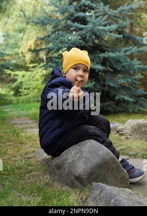Un bambino siede in un parco su una grande roccia e punta un dito direttamente a voi. Dito del ragazzo che indica la fotocamera. Primo piano verticale, messa a fuoco morbida. Foto Stock