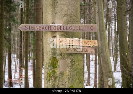 Strada in legno segno di percorso Steinbach-Quellgebiet, Quellenrundweg e Neunkirchen appeso su un albero durante l'inverno con neve sullo sfondo Foto Stock