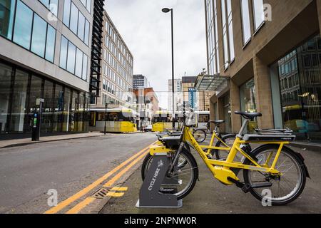 Biciclette gialle allineate su una strada a Manchester per il noleggio visto accanto a doppie linee gialle nel febbraio 2023 come due tram passa è lo sfondo. Foto Stock