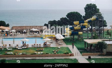 Offuscata dell'area dell'hotel con piscina e scivolo d'acqua durante la pioggia. I lettini sono vuoti, le persone fuggono dalla pioggia sotto la tettoia del Beach Cafe. Concetto Foto Stock